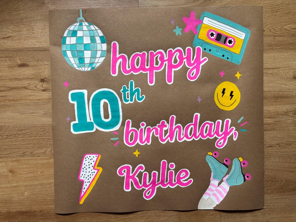 A brown cardboard birthday sign printed with 'happy 10th birthday, Kylie' in pink and teal lettering, decorated with retro-style illustrations including a disco ball, cassette tape, smiley face, lightning bolt, roller socks, and stars, photographed flat on a wooden floor.
