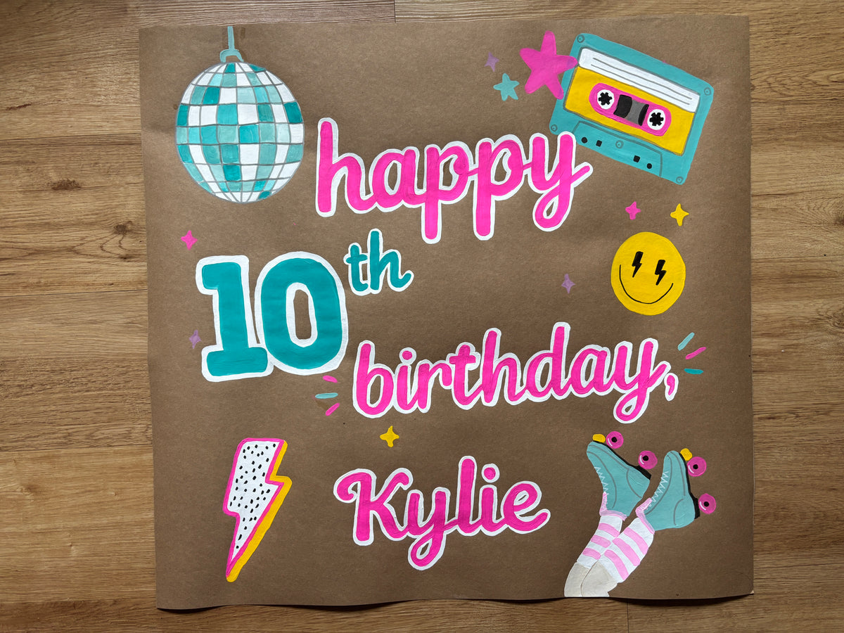 A brown cardboard birthday sign printed with 'happy 10th birthday, Kylie' in pink and teal lettering, decorated with retro-style illustrations including a disco ball, cassette tape, smiley face, lightning bolt, roller socks, and stars, photographed flat on a wooden floor.
