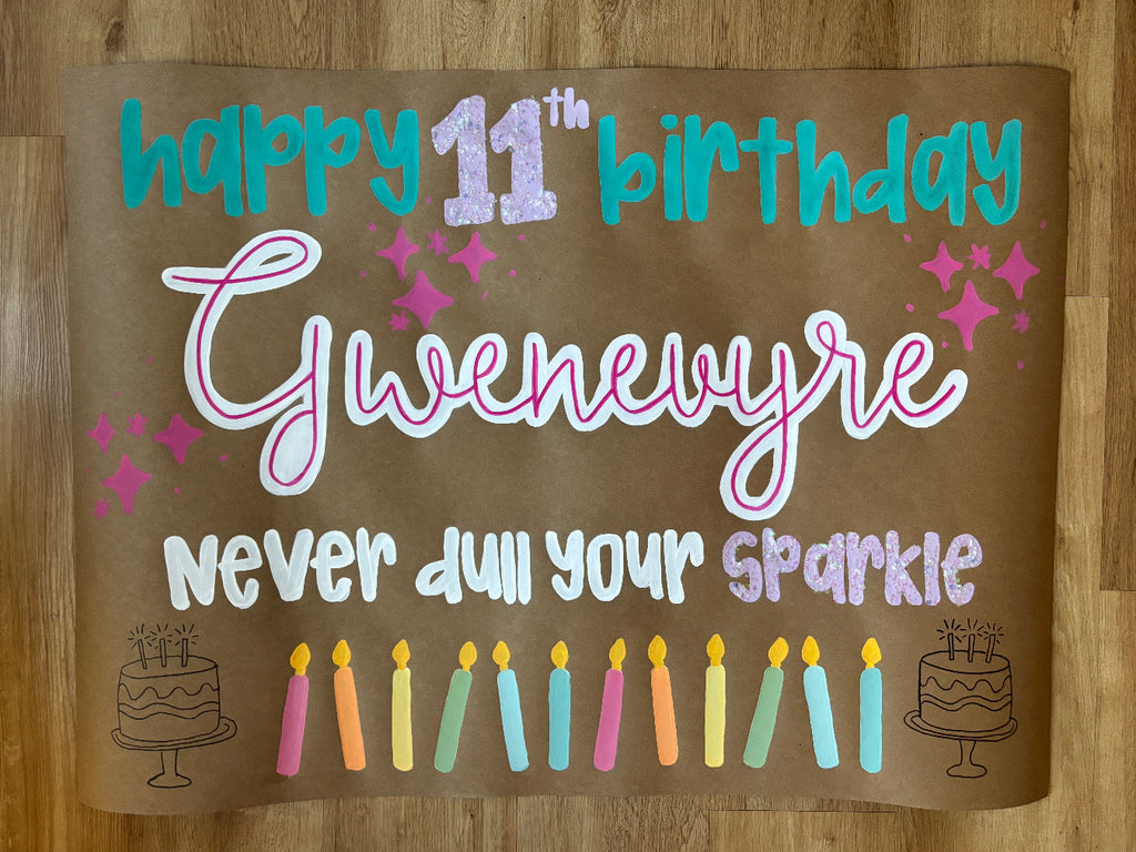 A custom 11th birthday banner or backdrop featuring the text 'happy 11th birthday Gwenwyre Never dull your sparkie' in colorful lettering on a brown background, decorated with candles, sparkles, and cake illustrations, laid flat on a wooden floor.
