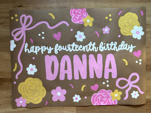 A square brown paper birthday card with colorful hand-painted floral designs and the text 'happy fourteenth birthday DANNA', photographed flat on a wooden floor.
