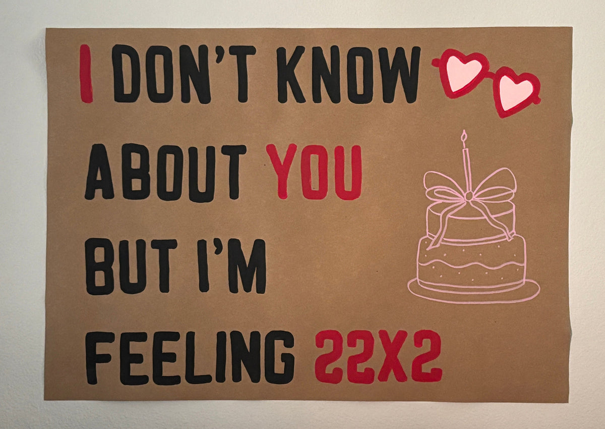 A square brown paper sign or poster with hand-lettered text reading 'I DON'T KNOW ABOUT YOU BUT I'M FEELING 22x2' in black and red, plus two pink hearts and a simple pink line drawing of a birthday cake. The sign is photographed against a plain light wall.
