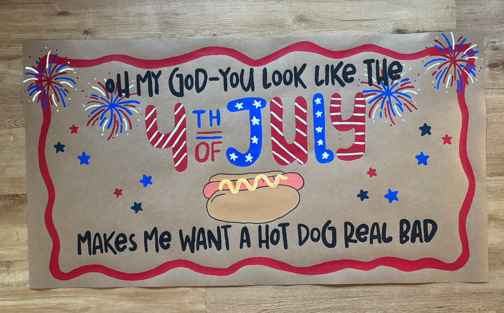 A rectangular patriotic sign with a brown paper-like background, red border, and hand-painted text celebrating 4th of July with fireworks, stars, and a hot dog illustration, placed on a wood surface.

