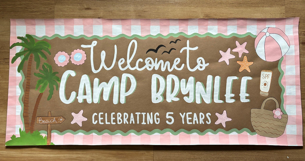 A rectangular printed banner laid flat on a wooden surface, featuring a pink-and-white gingham border, a brown center panel, and the text 'Welcome to CAMP BRYNLEE CELEBRATING 5 YEARS' with tropical and beach-themed illustrations including palm trees, flowers, stars, a beach ball, and a picnic basket.
