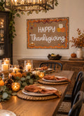 A rustic wooden dining room table set for Thanksgiving, decorated with candles, pinecones, pumpkins, and greenery, with a large 'Happy Thanksgiving' sign hanging on the wall behind.
