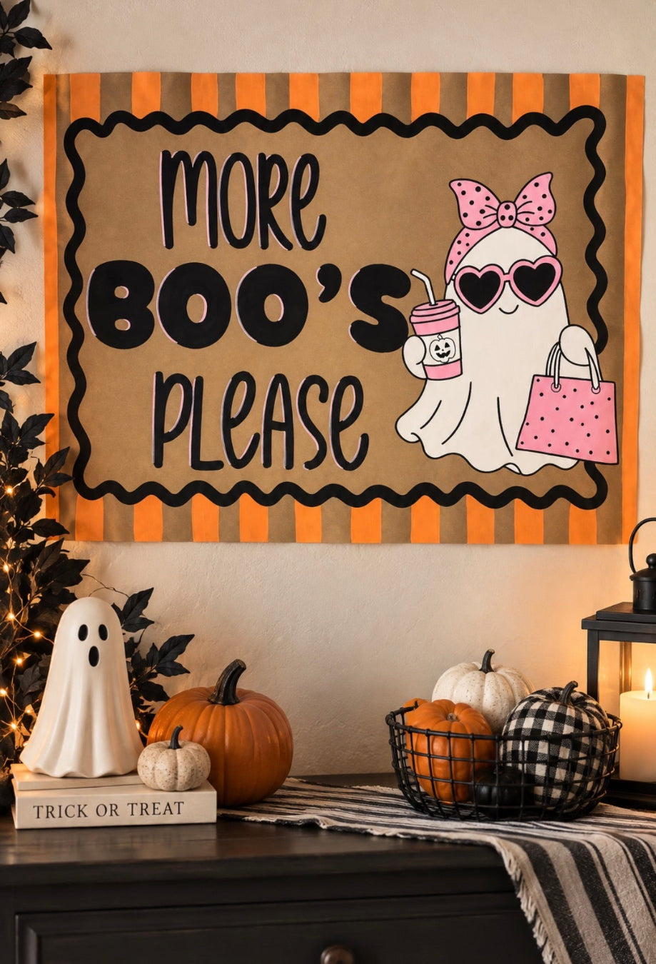 A Halloween-themed wall sign with the text 'MORE BOO'S PLEASE' in black lettering on a tan background, bordered by orange and black stripes. The sign features a cartoon ghost wearing a pink bow, heart-shaped sunglasses, and holding a pink polka-dot drink and bag.