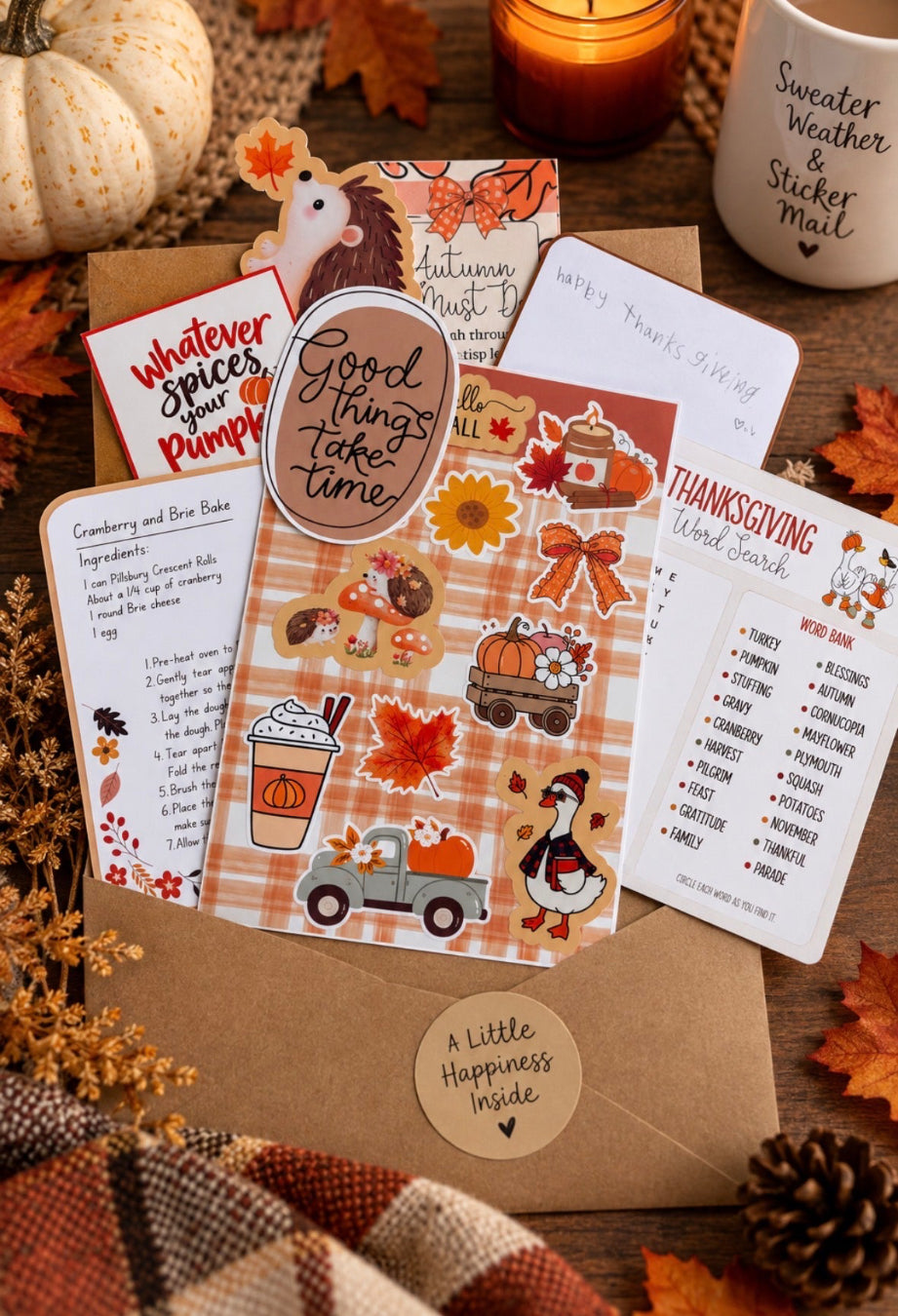 A styled flat-lay of autumn-themed stationery cards and stickers arranged on a kraft envelope with fall leaves, pumpkins, and a candle. The cards include Thanksgiving word search, cranberry and bacon bake recipe, and autumn muslin patterns, with colorful seasonal illustrations.
