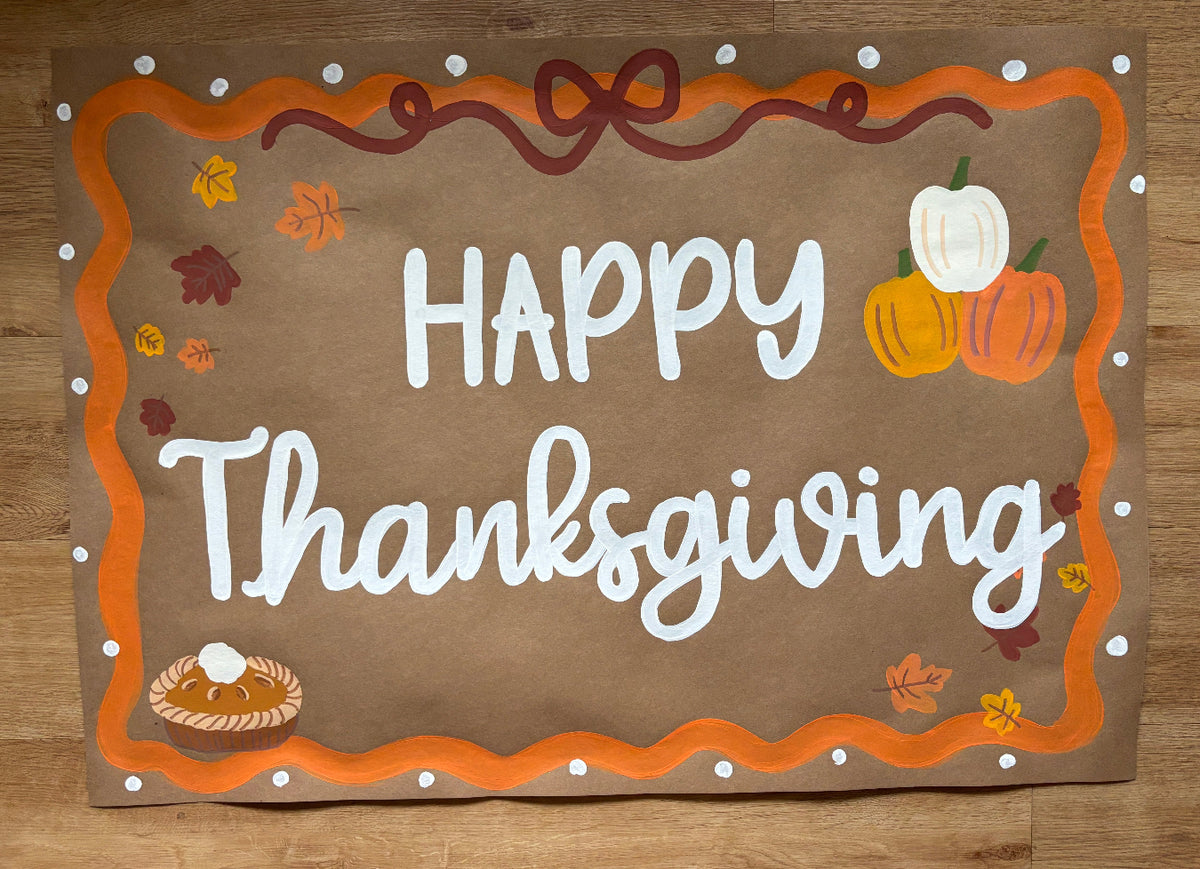 A handmade Thanksgiving-themed sign or placemat with 'HAPPY Thanksgiving' in white script on a brown background, surrounded by an orange scalloped border, white dots, autumn leaves, pumpkins, and a small pie illustration, photographed on a wooden surface.
