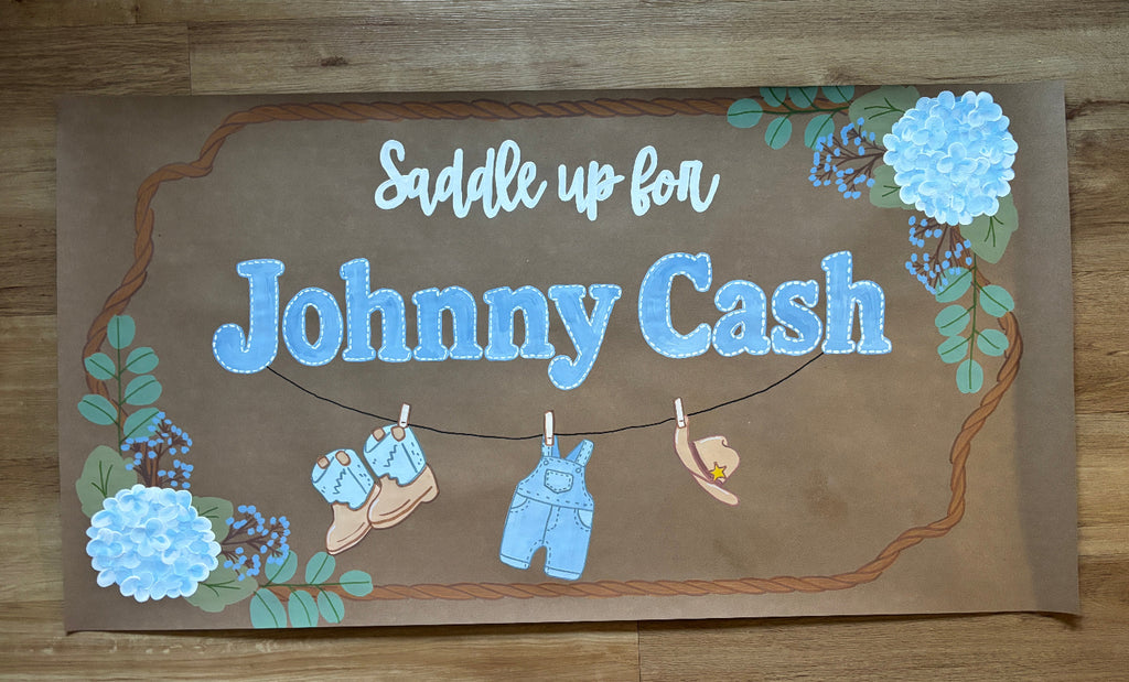 A rectangular brown sign with the words 'Saddle up for Johnny Cash' in white and blue lettering, decorated with blue flowers, green leaves, and small illustrations of baby shoes, overalls, and a hat hanging on a string, placed on a wooden floor.

