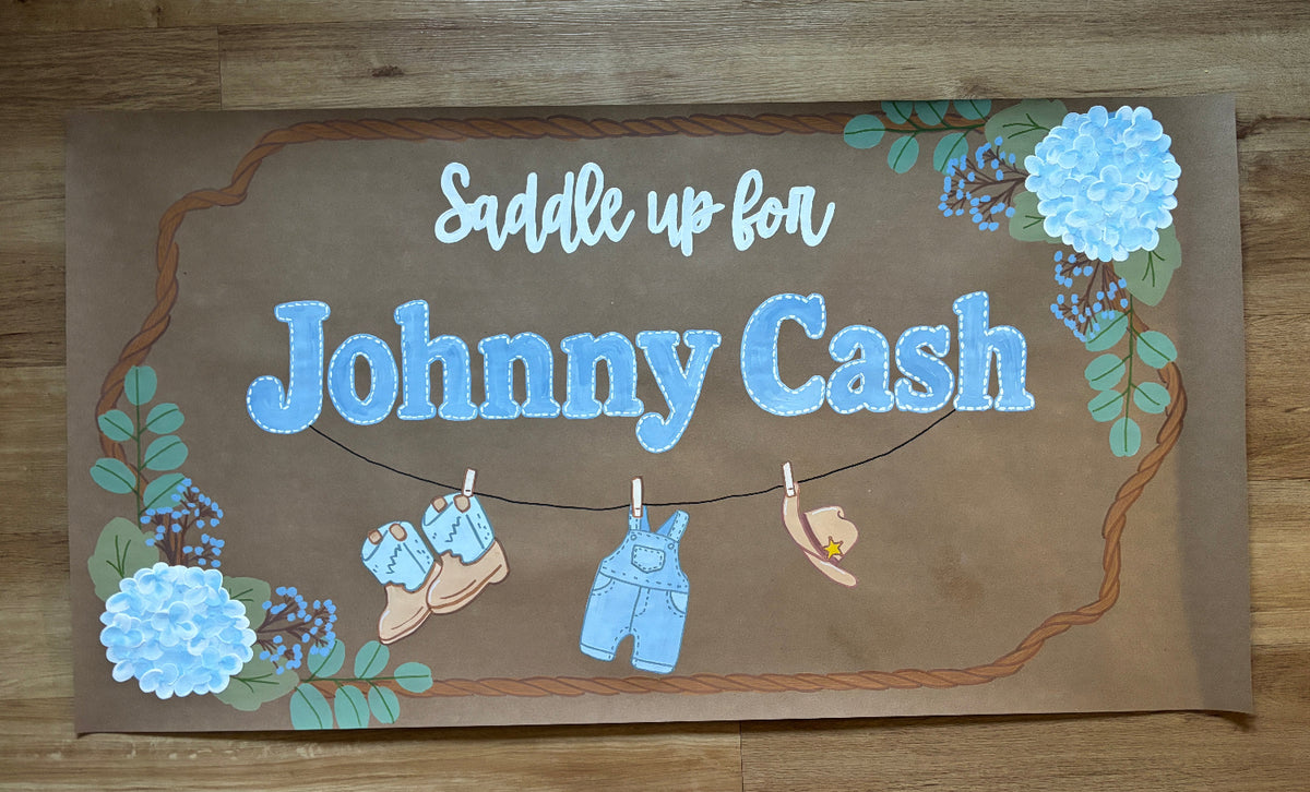 A rectangular brown sign with the words 'Saddle up for Johnny Cash' in white and blue lettering, decorated with blue flowers, green leaves, and small illustrations of baby shoes, overalls, and a hat hanging on a string, placed on a wooden floor.
