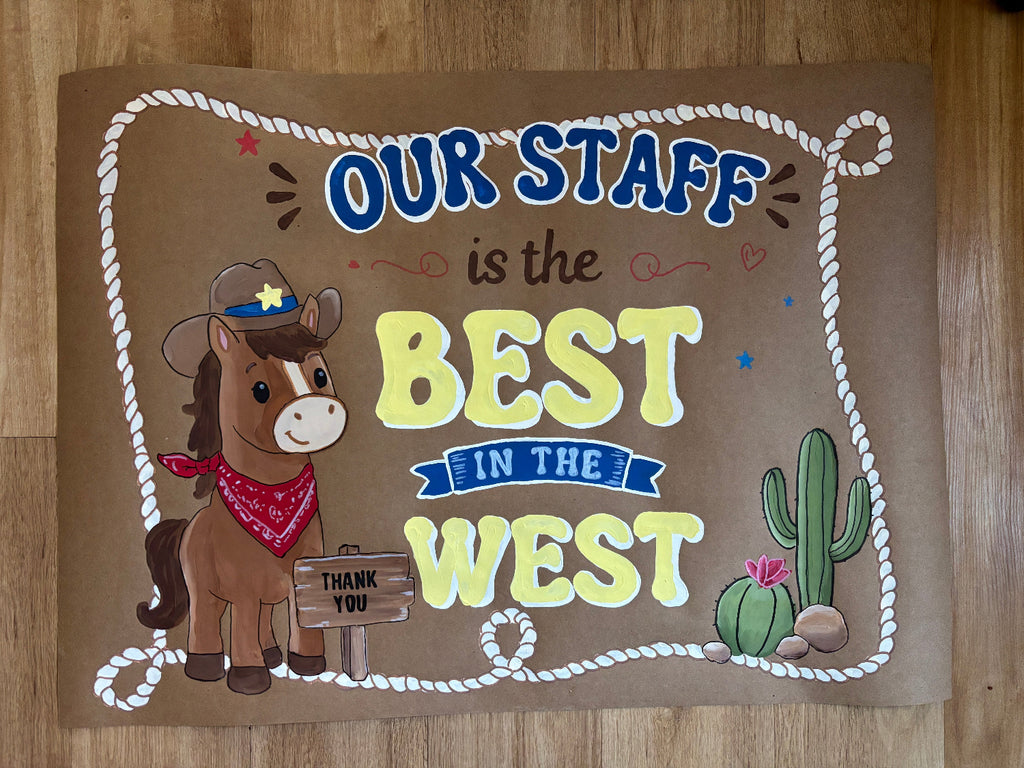 A rectangular brown sign with a western-themed design, featuring the text 'OUR STAFF is the BEST IN THE WEST' in blue and yellow lettering, a cartoon cowboy horse, a cactus, and a rope border, placed on a wooden floor.
