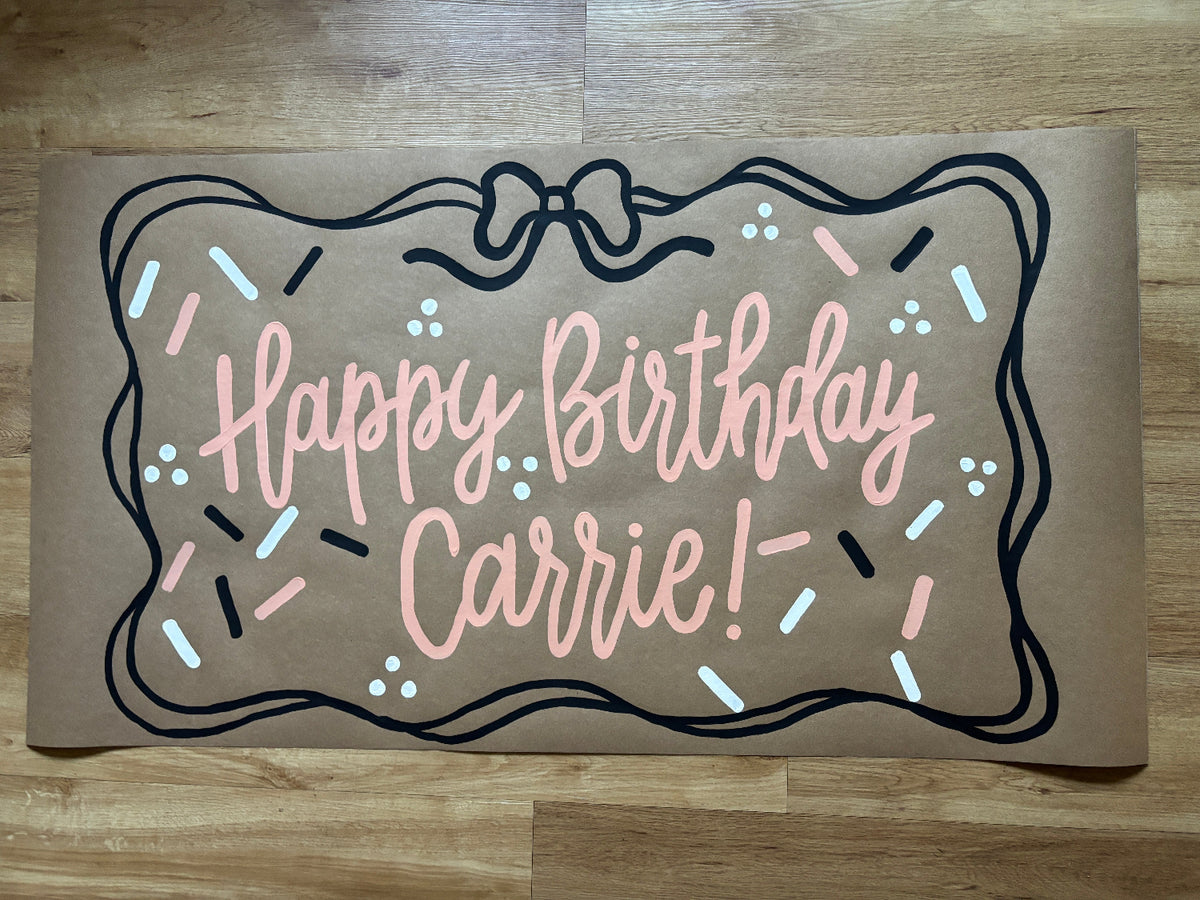 A brown kraft paper doormat with a hand-painted design that reads 'Happy Birthday Carrie!' in pink script, framed by a black outline with a bow and sprinkle details, shown on a wooden floor.
