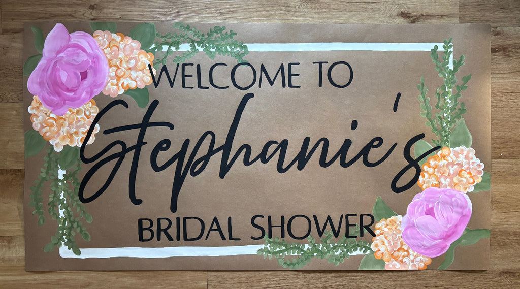 A rectangular wooden sign with a brown wood-grain finish, featuring the text 'WELCOME TO Stephanie's BRIDAL SHOWER' in black lettering, decorated with pink flowers, green leaves, and peach hydrangea-style clusters on a light wood background.
