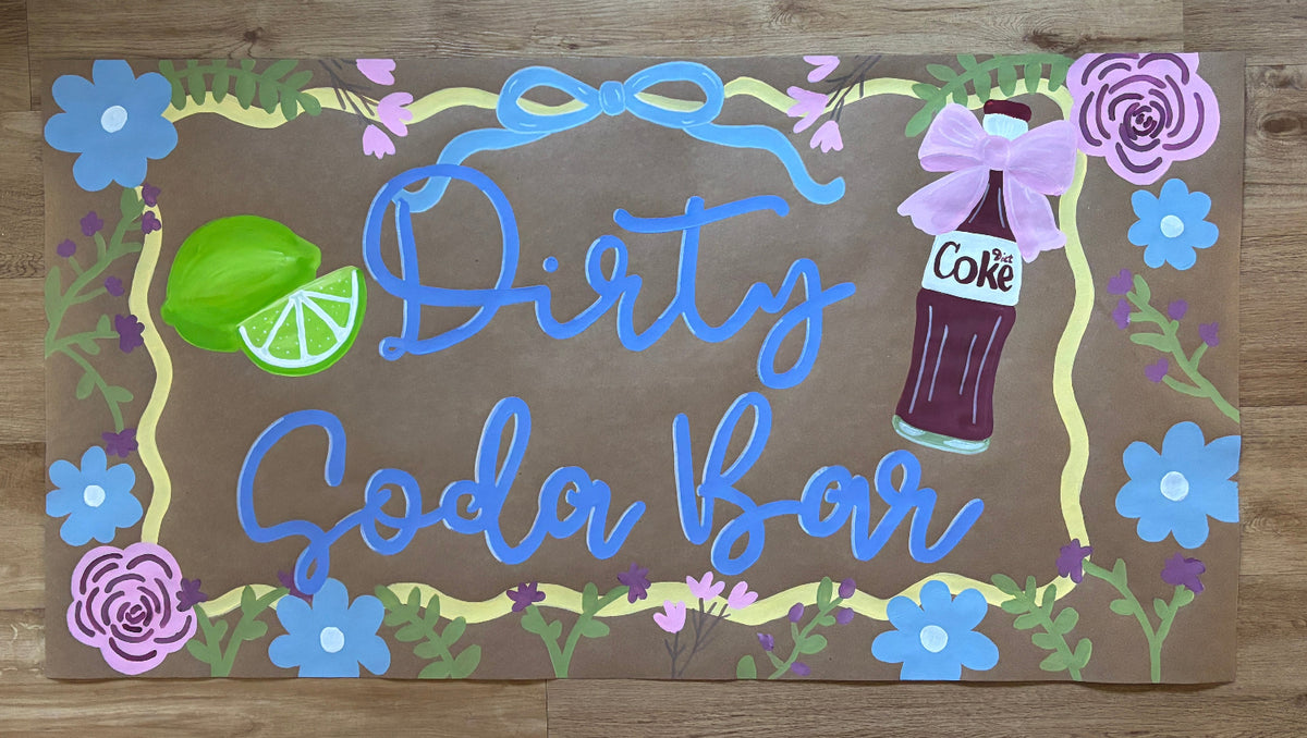 A rectangular decorative sign with the words 'Dirty Soda Bar' in blue script on a brown background, surrounded by painted flowers, green leaves, a lime slice, and a soda bottle illustration. The sign is photographed flat on a wood surface.
