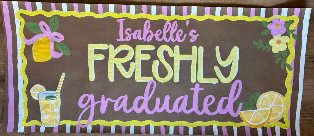 A custom rectangular graduation sign with a brown chalkboard-style background. The sign features the text 'Isabelle's FRESHLY graduated' in colorful hand-drawn lettering, decorated with illustrations of a yellow bell, a lemonade glass, and lemon slices, all framed by a wavy yellow border and pink, white, and yellow striped accents.