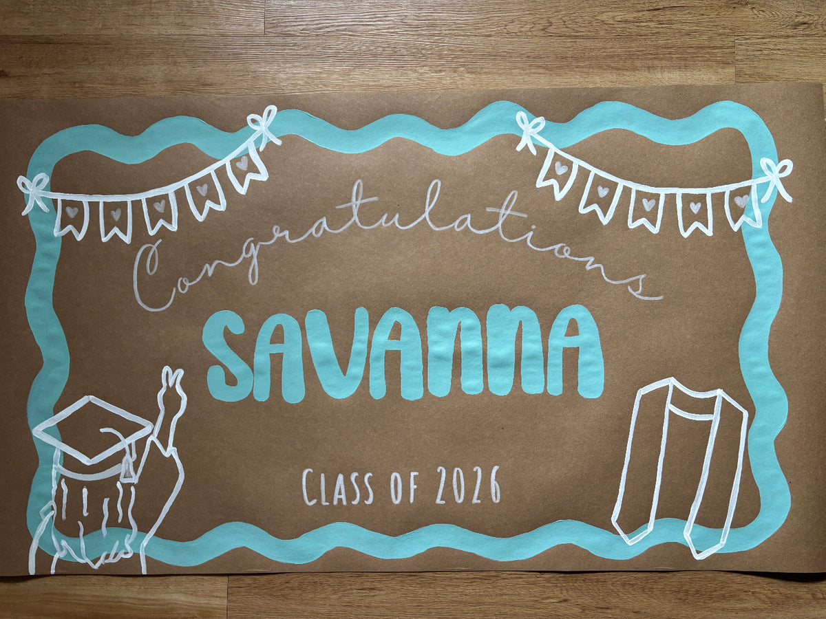 A custom graduation announcement board or sign with a brown background, hand-drawn light blue border, and white text reading 'Congratulations SAVANNA Class of 2026' along with graduation cap and gown illustrations, placed on a wooden floor.
