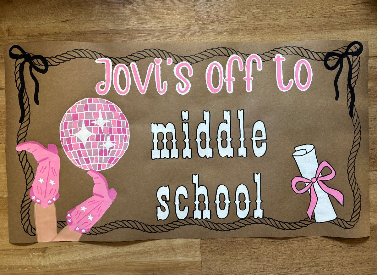 A brown cardboard sign with a rope border and the text 'Jovi's off to middle school' in pink and white lettering, decorated with a pink disco ball, pink-and-white gloves, and a rolled diploma, placed on a wooden floor.
