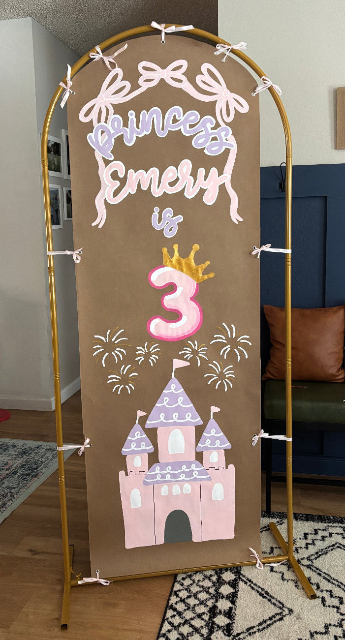 A freestanding arch-shaped banner stand in a home setting, featuring a printed birthday sign that reads 'Princess Emily is 3' with a pink number 3, white script text, fireworks, and a pink castle illustration on a brown background.
