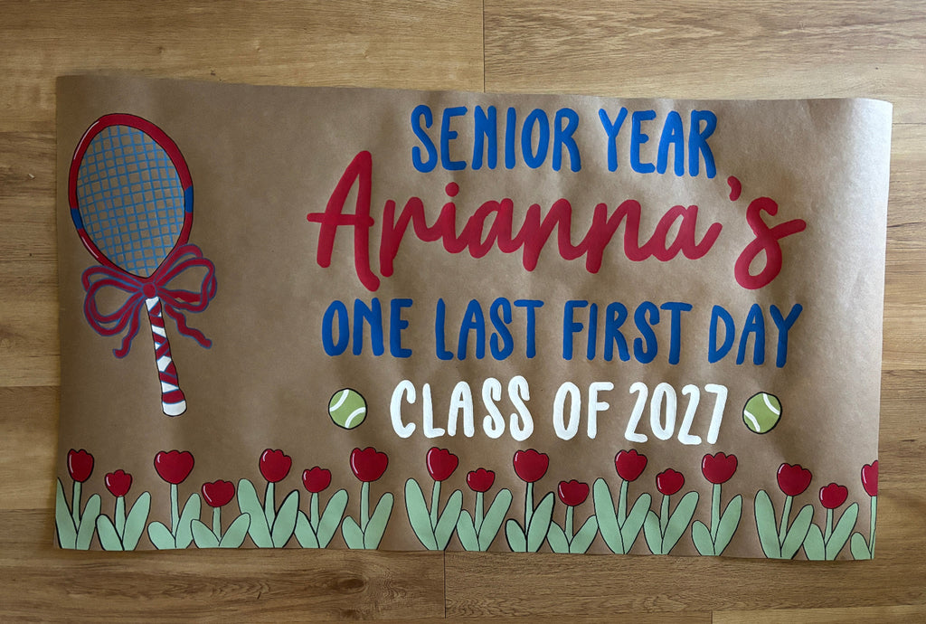 A personalized brown kraft paper banner for a tennis club's 'Senior Year' and 'Class of 2027' laid flat on a wooden floor. The banner features a red tennis racket, red tulips, green leaves, and blue and red lettering.
