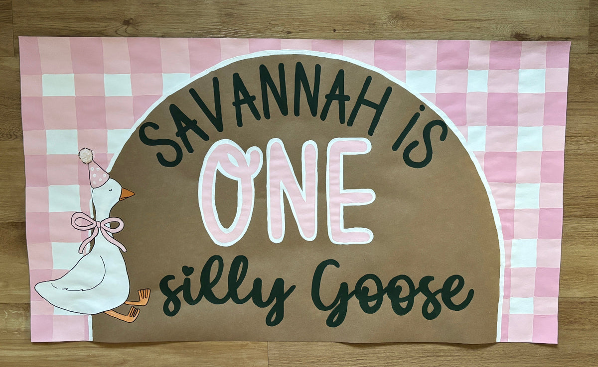 A custom fabric banner or flag with a pink and white gingham border, featuring a brown central arch with the text 'SAVANNAH IS ONE silly Goose' in dark green and white lettering, alongside an illustration of a white goose wearing a party hat and bow.
