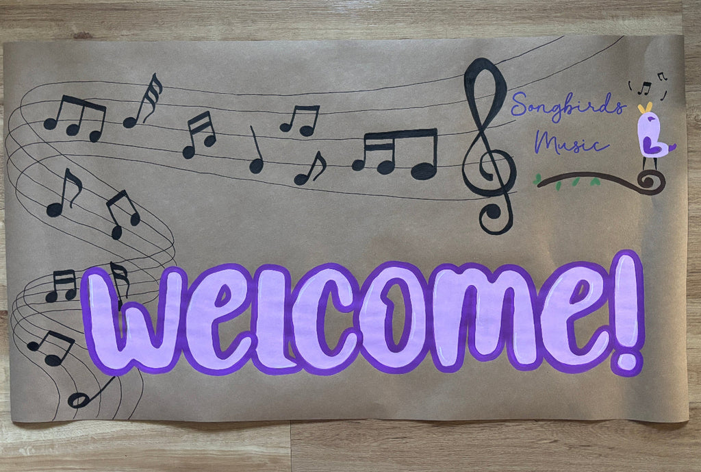 A rectangular brown paper sign with a music theme, featuring musical notes on staff lines, the text 'Songbirds Music' in purple and green, and a large purple 'Welcome!' across the bottom, placed on a light wood floor.
