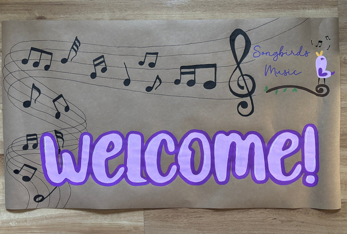 A rectangular brown paper sign with a music theme, featuring musical notes on staff lines, the text 'Songbirds Music' in purple and green, and a large purple 'Welcome!' across the bottom, placed on a light wood floor.
