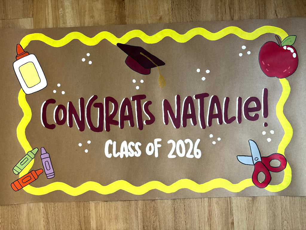 A handmade graduation banner for 'Natalie Class of 2026' decorated with school-themed cutouts including a graduation cap, apple, scissors, crayons, and glue bottle on a wood floor background.
