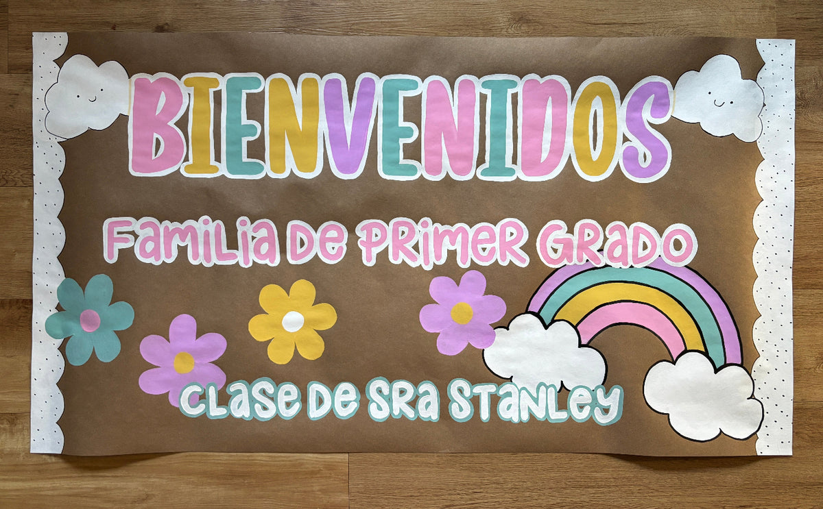 A handmade brown welcome sign for a first-grade class, featuring colorful hand-lettered text, paper flowers, clouds, and a rainbow, displayed flat on a wooden floor.
