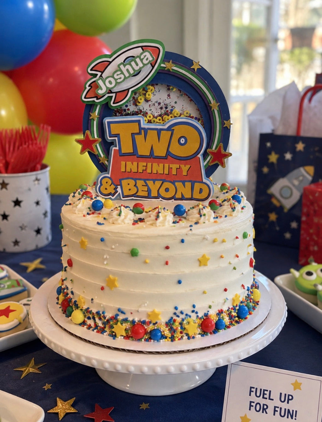 A white frosted birthday cake with colorful sprinkles and a 'Joshua Two Infinity & Beyond' topper, displayed on a white cake stand at a party table with balloons and party decorations in the background.
